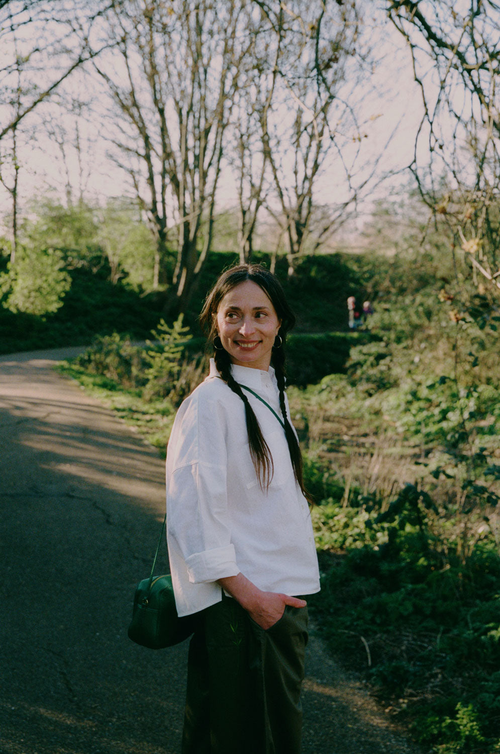 A woman is standing on a path in a green park surrounded by nature. She is turned to the side looking over her shoulder, smiling, with her hand in her trouser pocket. She wears Saywood's white recycled cotton Lela Boxy Shirt with the khaki Amelia Wide Leg Trousers. Her hair is plaited in pigtails and she has a green cross body bag across her.