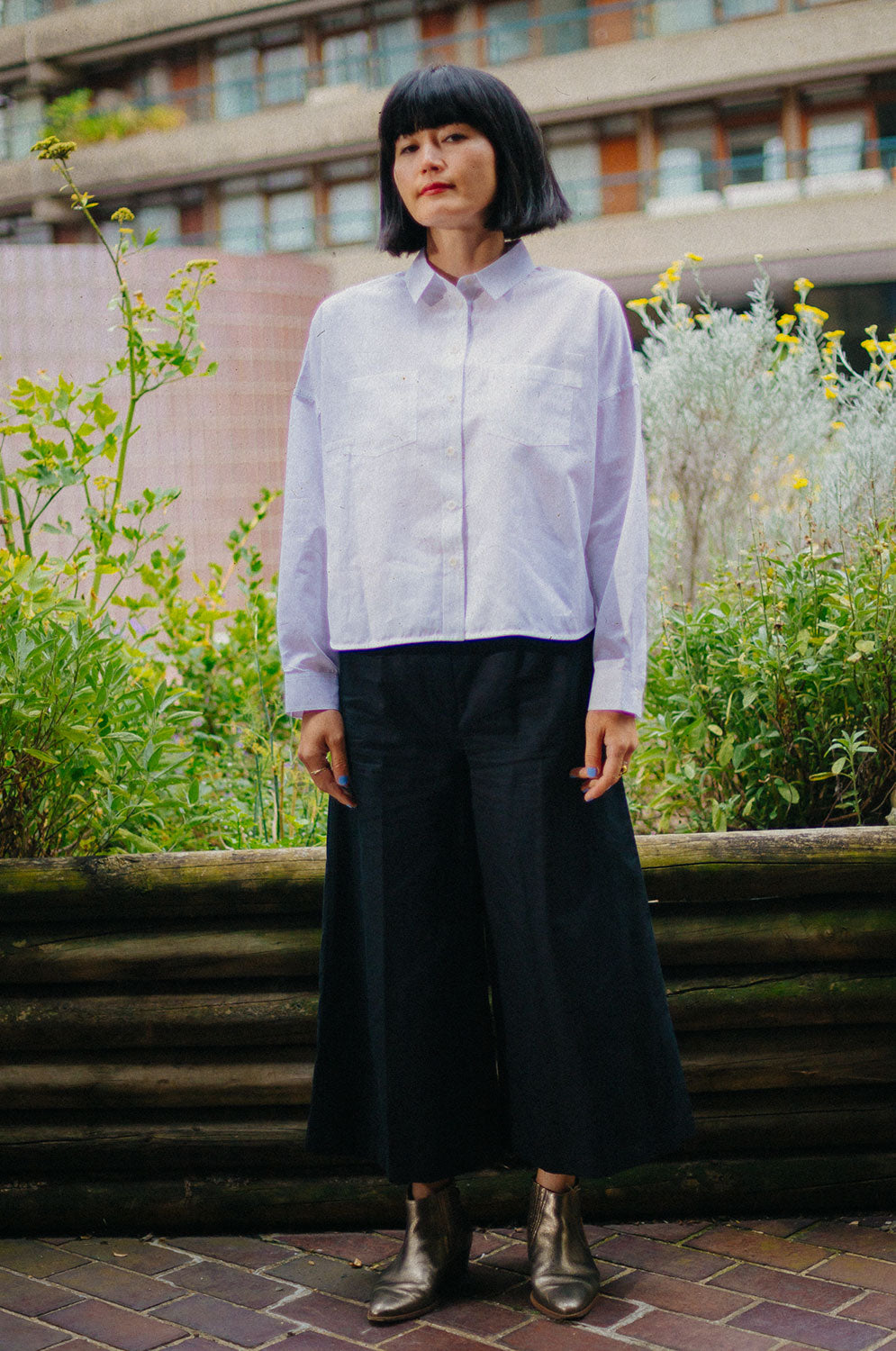 A woman stands looking at the camera with hands by her side. She is wearing Saywood's white recycled cotton Lela Boxy Shirt with patch pockets on the chest, and the black Amelia Wide Leg Culotte Trousers with gold ankle boots. Behind her is a green and yellow plant in a wooden trough, and the flats of the Barbican Centre can be seen in the background.