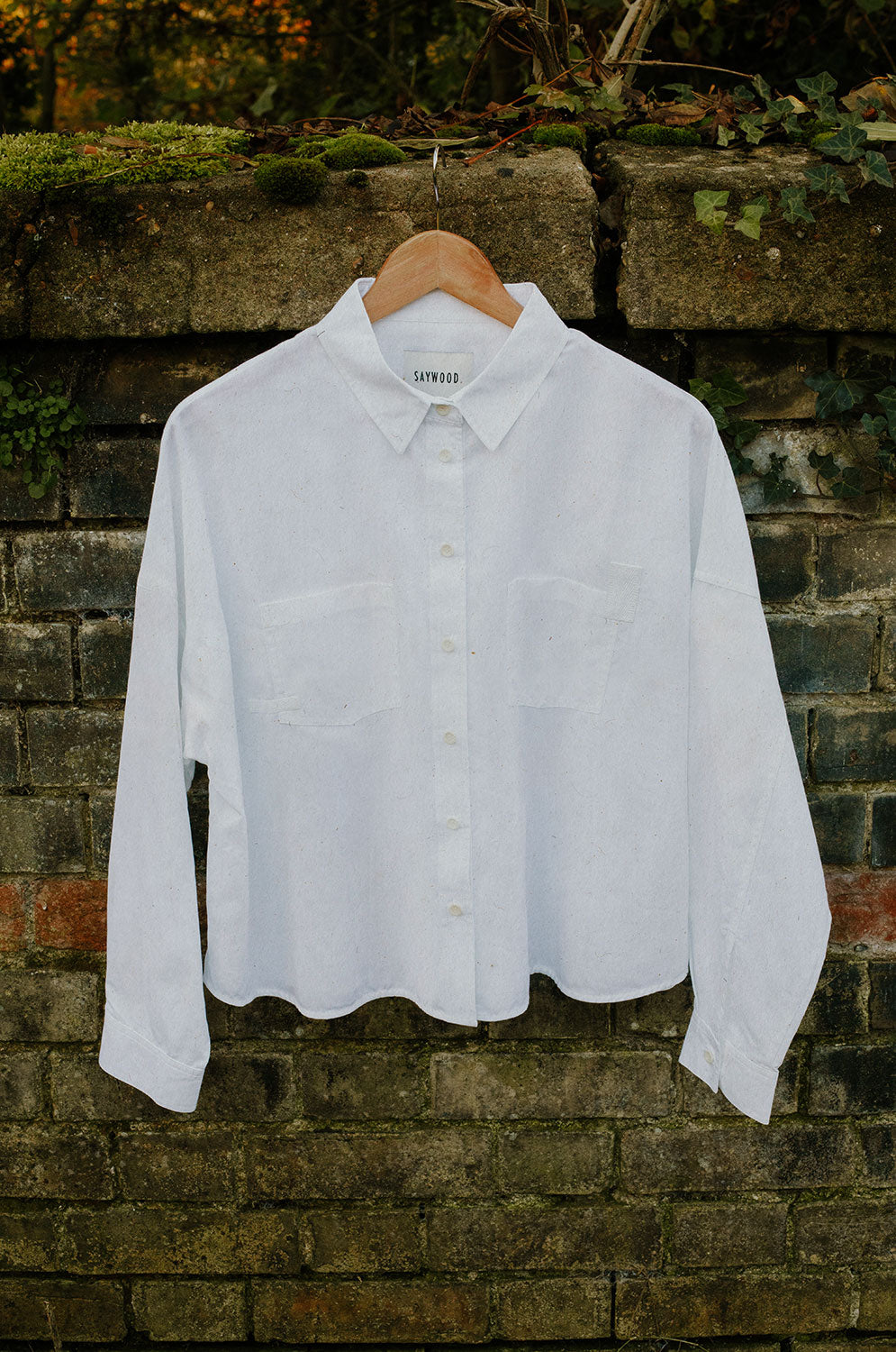 Women's white recycled cotton boxy shirt hanging on a hanger on a brick wall with moss scattered on top of the wall - The Lela Shirt, with patch pockets and cotton herringbone tape detail on the pocket corners. Made in the UK. By Saywood