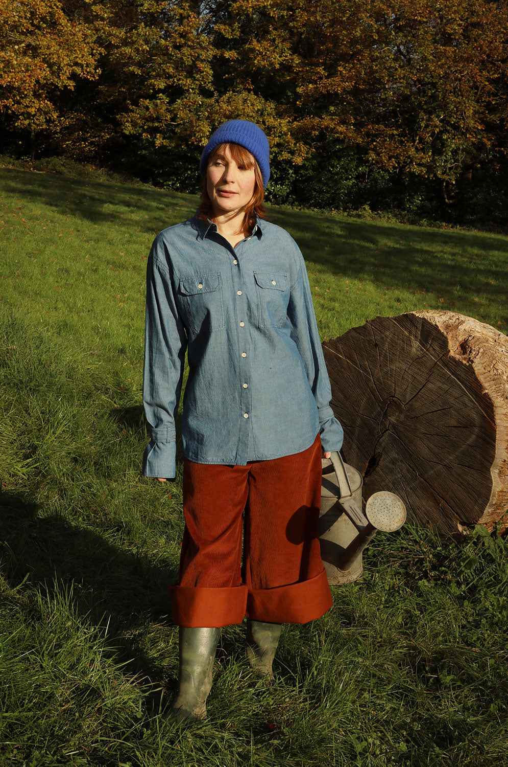 A woman standing in a green field with trees behind her and a big tree trunk in the background. She is wearing Saywood's Caramel Tan Corduroy Trousers in Organic Cotton, styled rolled up with wellies, with the cotton Zadie Boyfriend Shirt in Japanese Denim, and a blue beanie hat. She holds a watering can.