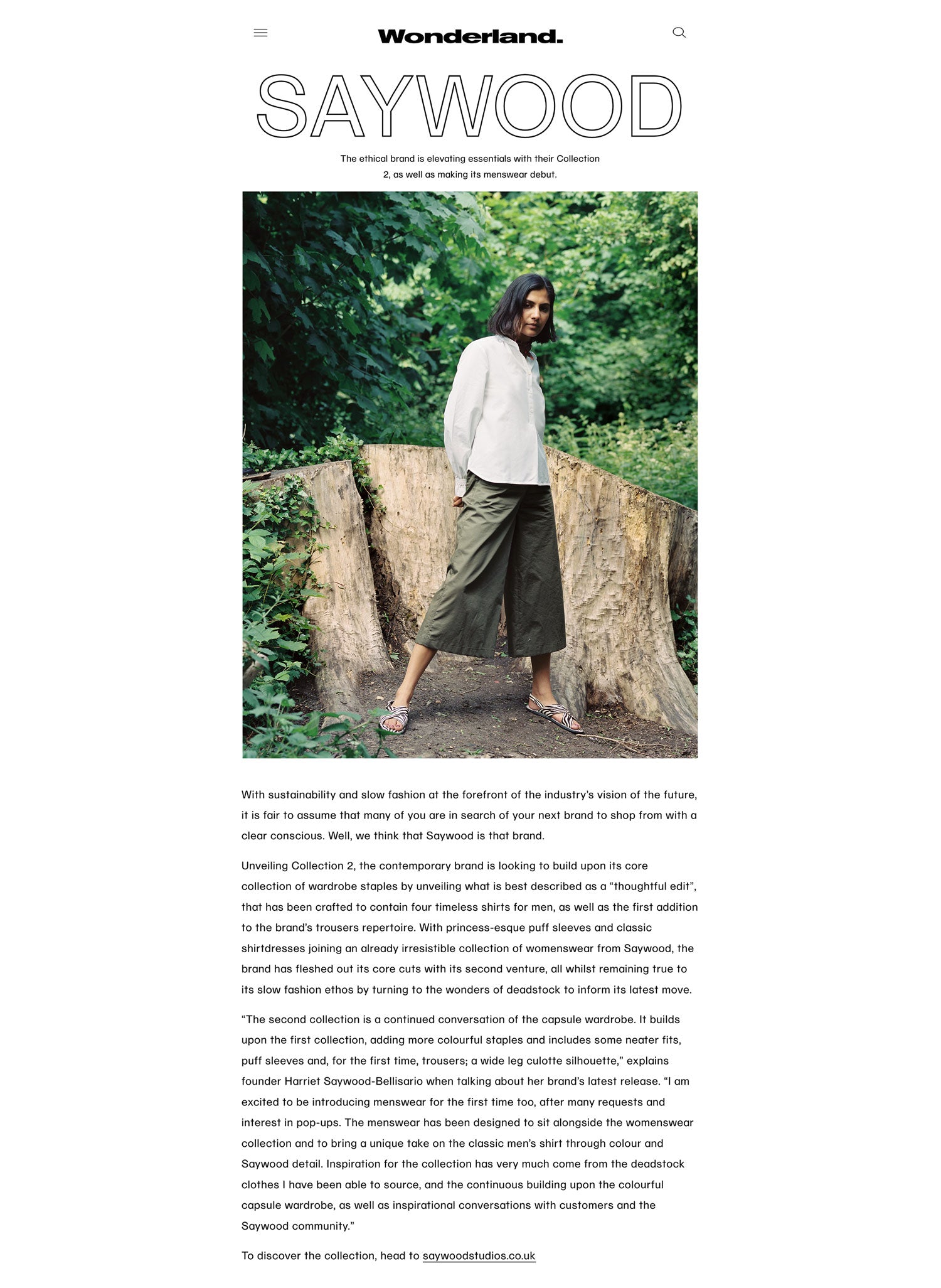 Image shows Saywood feature from Wonderland Digital. Text above and below image about the brand, with an image showing model wearing the Marie Blouse in white recycled cotton and the khaki Amelia Trouser, standing in front of green foliage and tree stumps
