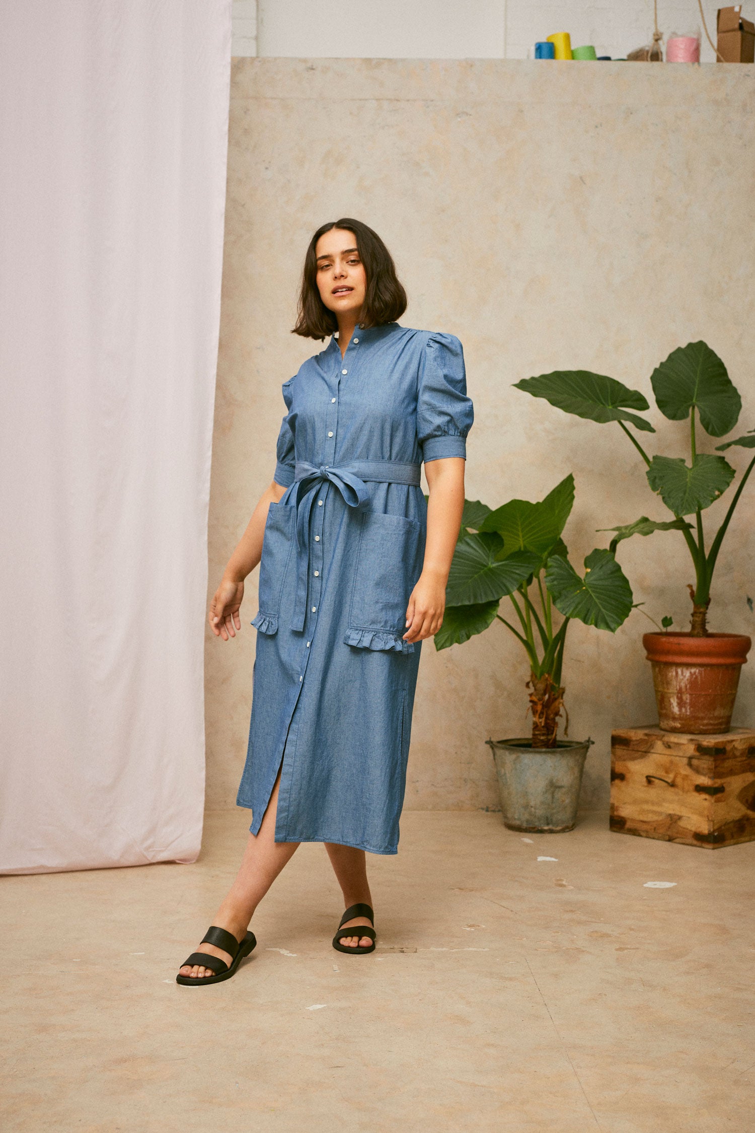 Full length shot of model wearing Saywood's Rosa denim shirtdress in light wash Japanese denim; a long line style with patch pockets and ruffles, a belt tied round the waist. Worn with black sandals. A plant and drop of pink fabric can be seen in the background.