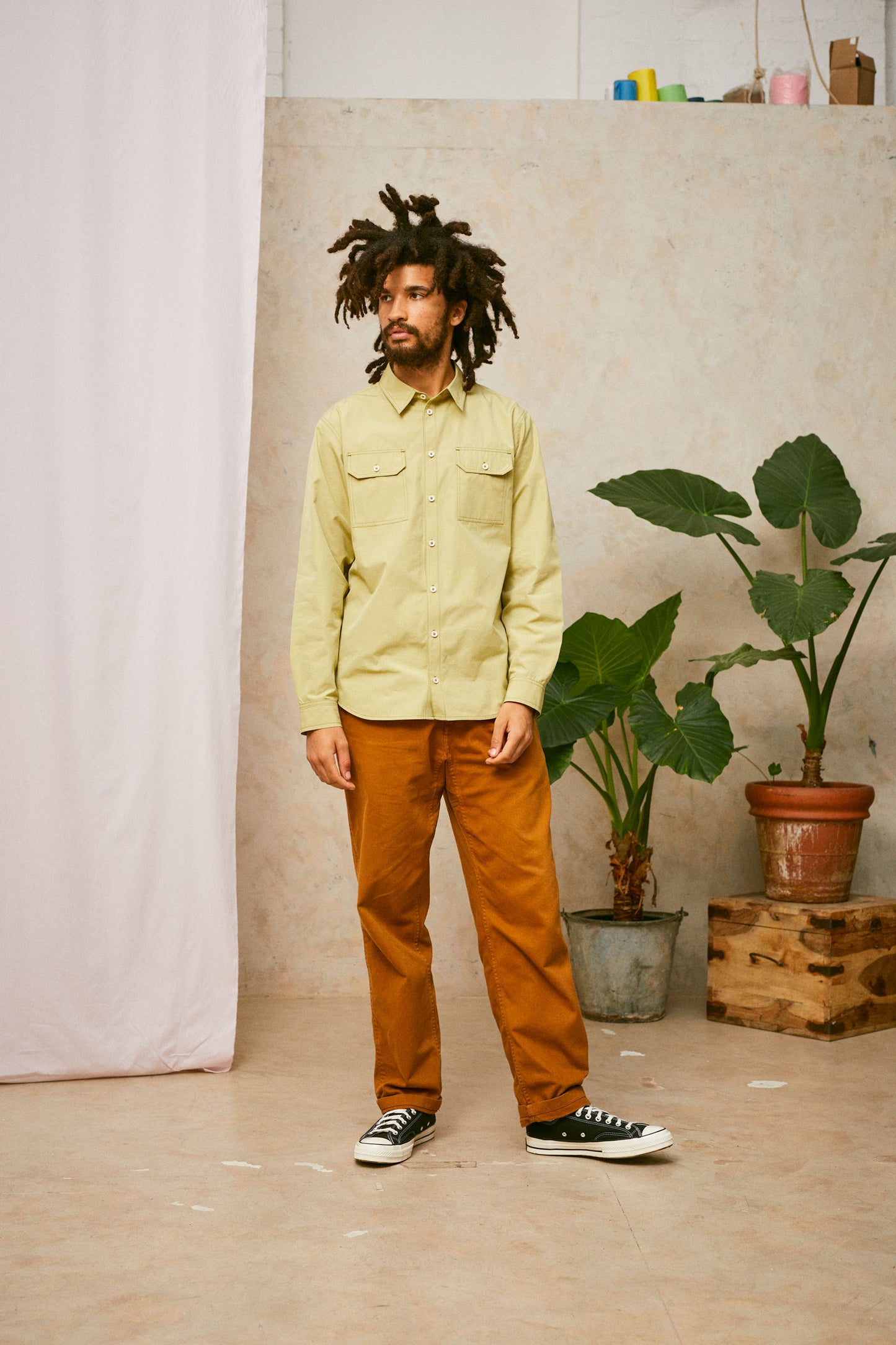 Full length shot of model wearing Saywood's Eddy Mens Khaki Shirt with utility pockets. Worn with tabacco trousers and black Converse. A plant and drop of pink fabric can be seen in the background.