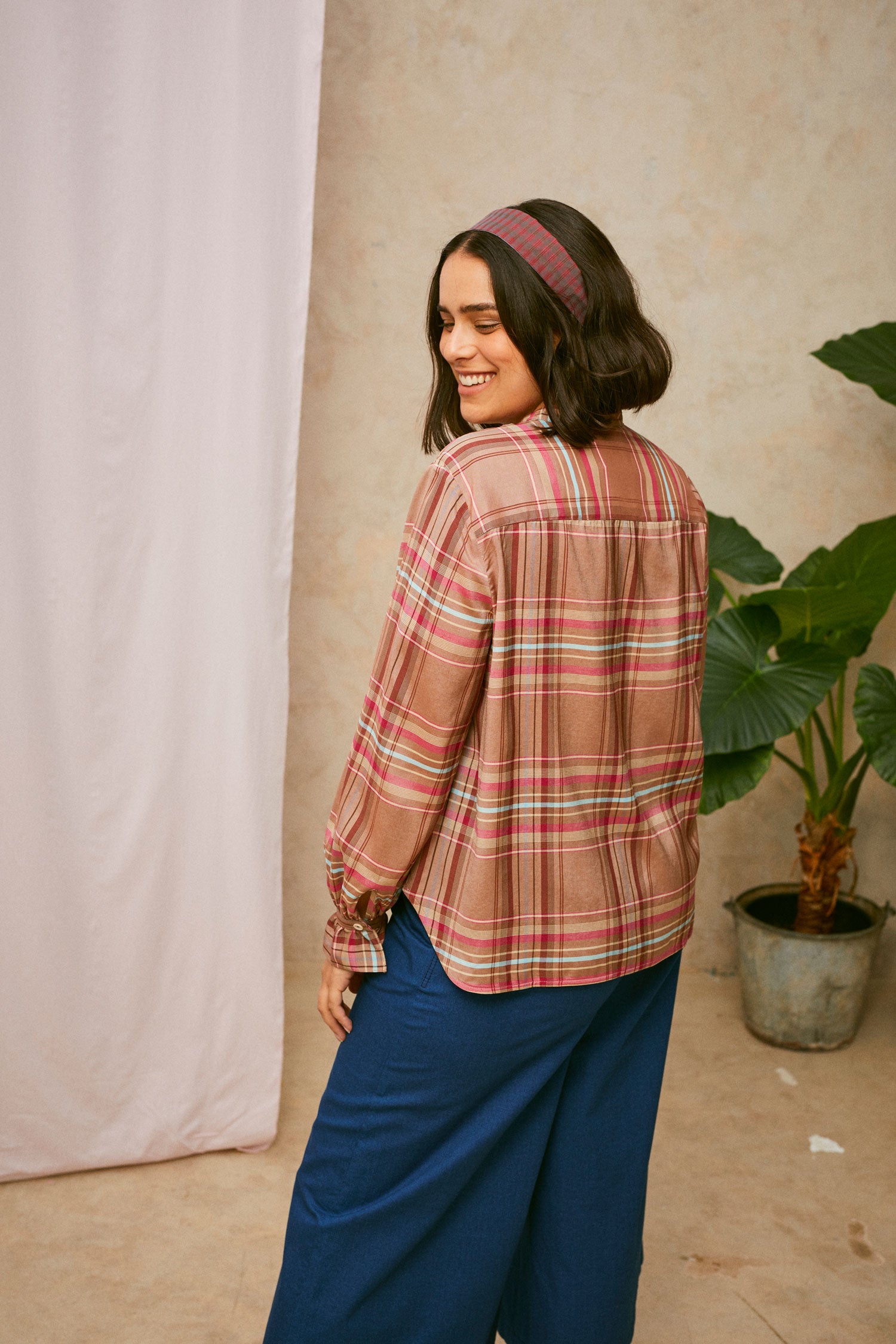 Model stands with her back to the camera. She wears Saywood's pink check shirt, the Marie A-Line Blouse, with natural indigo Amelia wide leg trousers. The red check Heidi Headband is worn in her her. A plant and drop of pink fabric can be seen in the background. 