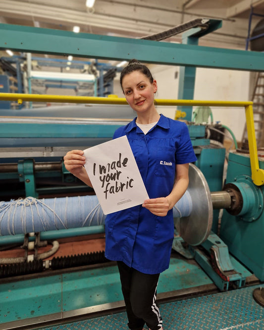 Fashion Revolution Week: Fabric maker and warper, Guilia, stands in front of a fabric machine in the fabric mill, Canclini in Italy, wearing a blue coat, holding a sign saying 'I made your fabric'.