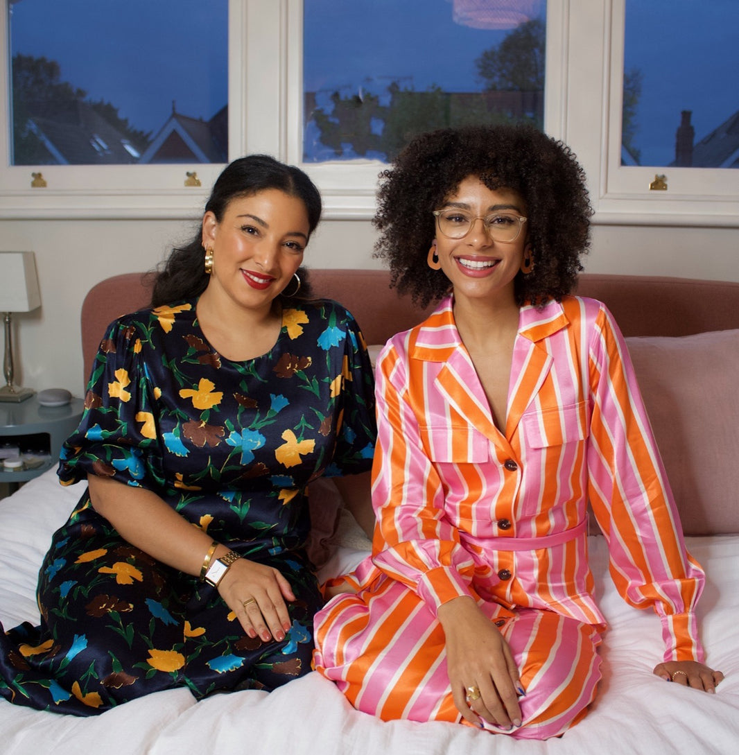 Sustainably Influenced Podcast co-founders and influencers Bianca F Foley and Charlotte Williams.  Bianca wears a black dress with a blue, ocre and chocolate print. Charlotte wears a pink an orange bold stripe pyjama set. They both are sitting on a bed.