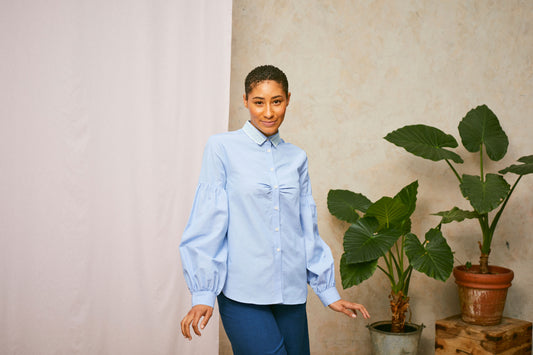 Model wears Saywood's Edi lace collar shirt with volume sleeves in pale blue recycled cotton. Pink fabric hangs in the background with two plants to the right. Her arms are stretched downwards with her hands flicking up slightly.