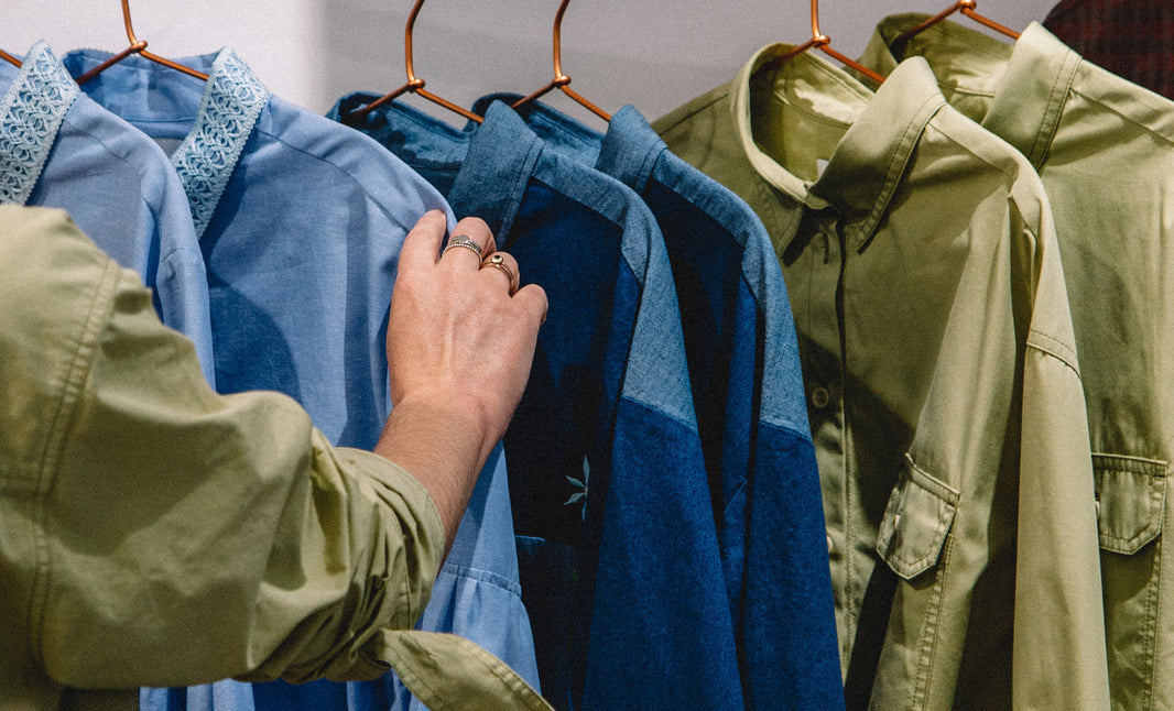 Beautiful blue and green Saywood shirts hanging on a rail, with a hand outstretched touching one of the garments. The woman is wearing the Saywood olive green Zadie Boyfriend Fit Shirt, and this can be seen on the arm.