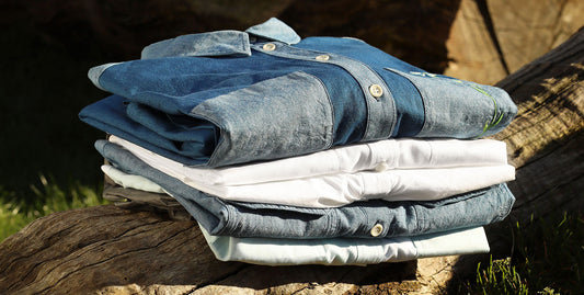 Saywood shirts folded in a pile on top of a log. White shirts, Japanese Denim shirts and shirtdress, green Lela Shirt