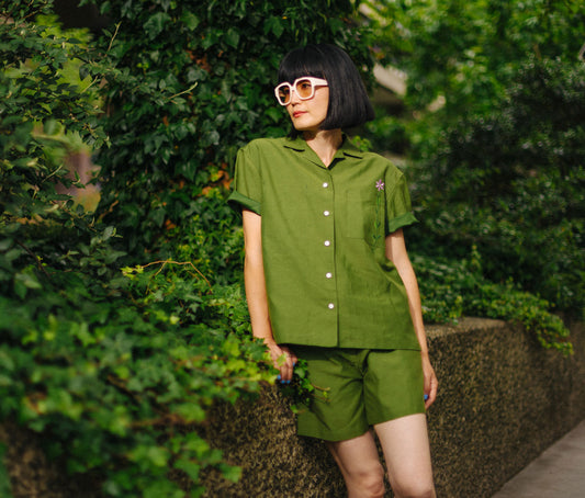 Women leans against a concrete wall, surrounded by green ivy. She is wearing sunglasses, worn with Saywood's matching co-ord set; Frances Unisex Cuban Shirt and Bessie A-Line Shorts in olive green, both made in the UK