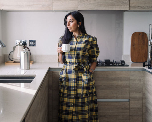 Female entrepreneur and founder Leena Parmar stands in her kitchen with a cup of coffee. She is wearing Saywood's Rosa Puff Sleeve midi Shirtdress in yellow check