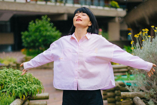 Woman stands amongst flower beds with her arms outstretched, looking up to the sky with her eyes closed. She is wearing Saywood's pink and orange boxy fit Lela Shirt, with black organic cotton and linen Amelia Trousers, made in the UK