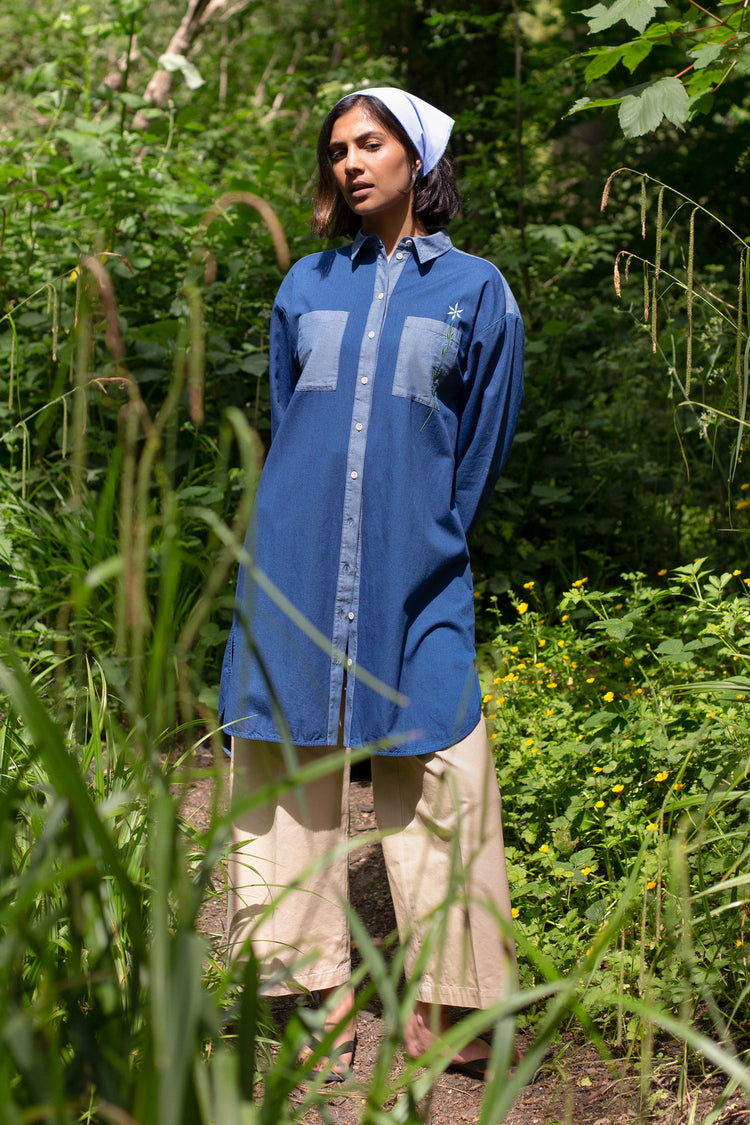 Model stands, surrounded by wild plants and greenery, with her hands behind her back. She wears Saywood's Etta Oversized Shirtdress in Japanese denim, worn over beige trousers, with a pale blue bandana in her hair made from recycled cotton.