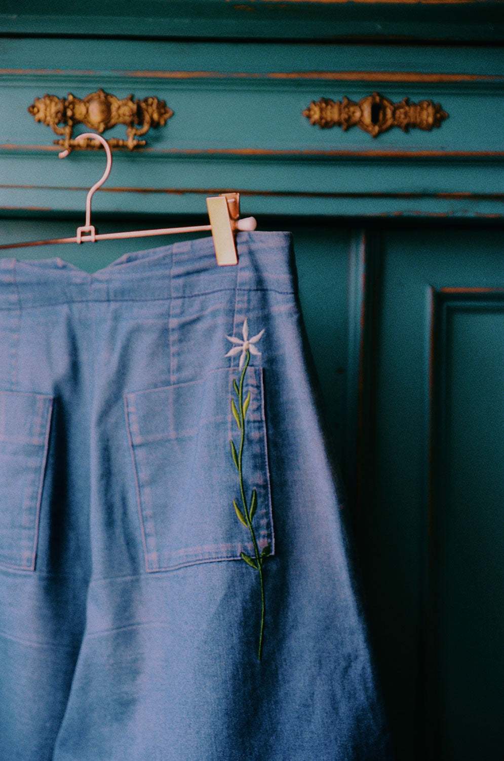 Saywood's Amelia Wide Leg Trousers in indigo Japanese Denim with signature embroidered flower seen on the back pocket, hangs on a rustic turquoise wardrobe, with metal cabinet above.