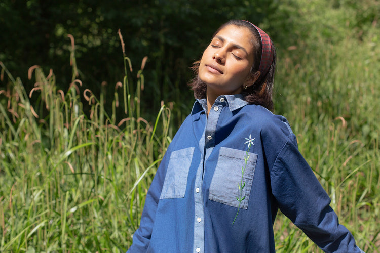 Model leans back with one arm behind her, surrounded by greenery and nature. She wears Saywood's Etta Oversized Shirtdress in Japanese denim, th embroidered flower on the chest pocket.