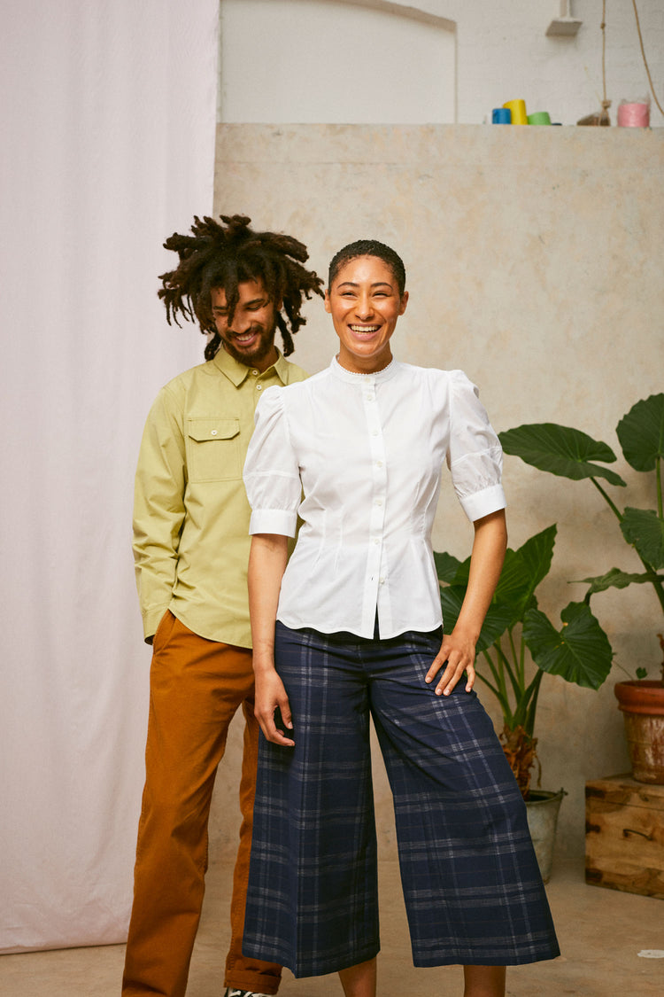 Male and female model stand together laughing, with plants and pink fabric in the background. He wears Saywood's khaki Eddy Shirt and she wears Saywood's puff sleeve blouse in white with navy check trousers.