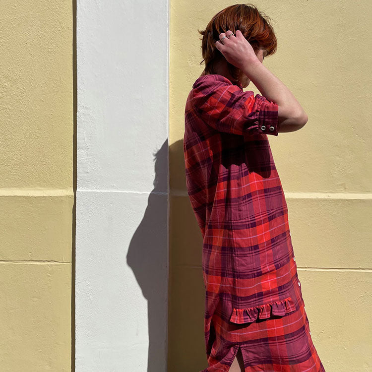 Woman leans against a concrete wall, surrounded by green ivy. She wears ivory sunglasses and Saywood's Frances Unisex Cuban Shirt in olive green, and the Bessie A-line Shorts in matching olive green, both made in the UK.