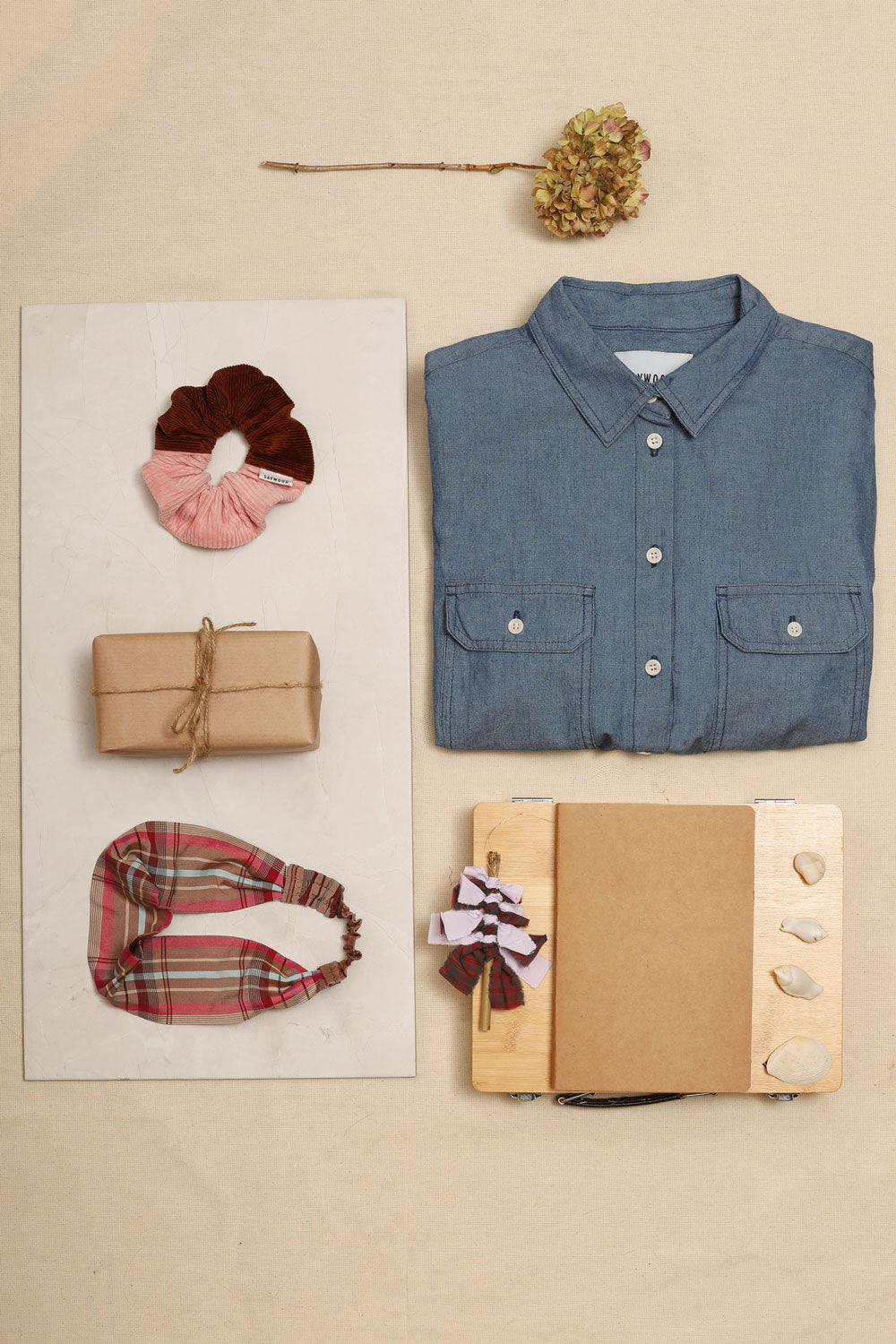 Luxury sustainable gifts for her. Laid our on an ivory background, folded Japanese Denim Zadie Shirt, pink and caramel corduroy scrunchie, pink check headband lilac and red tartan Christmas decoration, with a wrapped present, notebook, and dried flower.