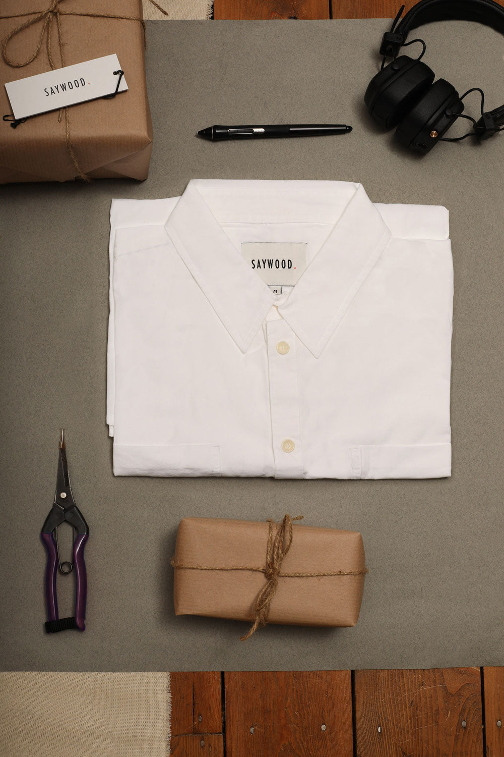 laid on a grey background on wooden floorboards, Saywood's mens' white shirt with pockets, made in london. With a pair of headphones, a pen, garden snips, and wrapped presents.
