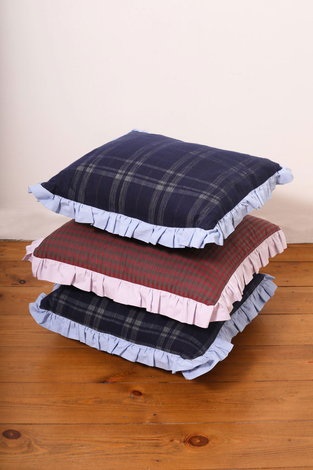 Three sustainable ruffle cushions, made in London, stacked on top of each other on a wooden floor with a white wall background. 2x navy tartan with a pale blue ruffle, 1x red mini check with a lilac ruffle. British wool fillers.