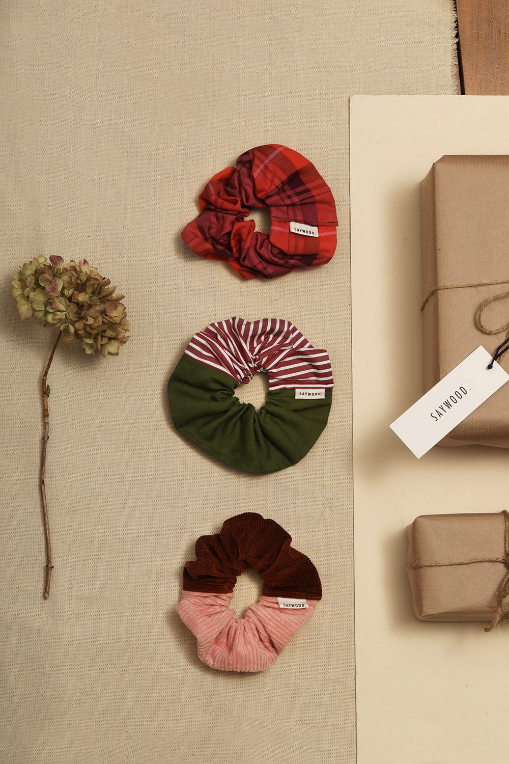 Sustainable stocking fillers. Laid on a cream background, three scrunchies; red merlot coral tartan scrunchie, green and red and white stripe scrunchie, pink and caramel corduroy scrunchie, with wrapped presents and a dried flower.