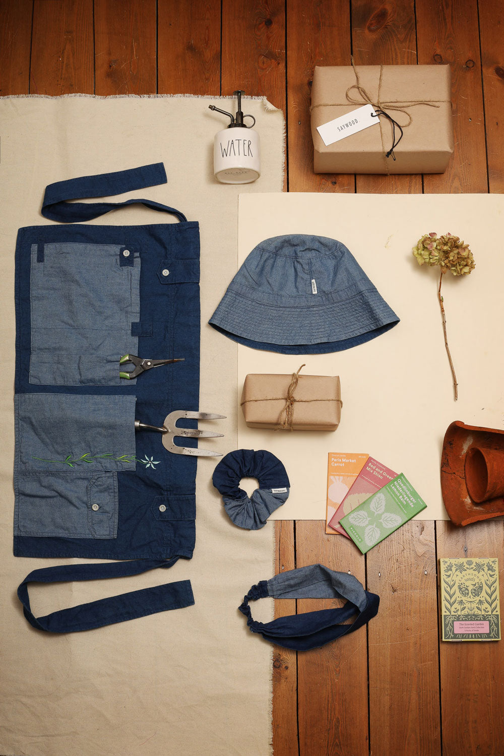 Laid on ivory cloth on wooden floor boards, a selection of gardening tools and gifts. Saywood's Japanese denim Multifunctional Gardening Tool Belt, Denim Reversible Bucket Hat, Patchwork Scrunchie in Japanese Denim, matching Thandi Twisted Headband.