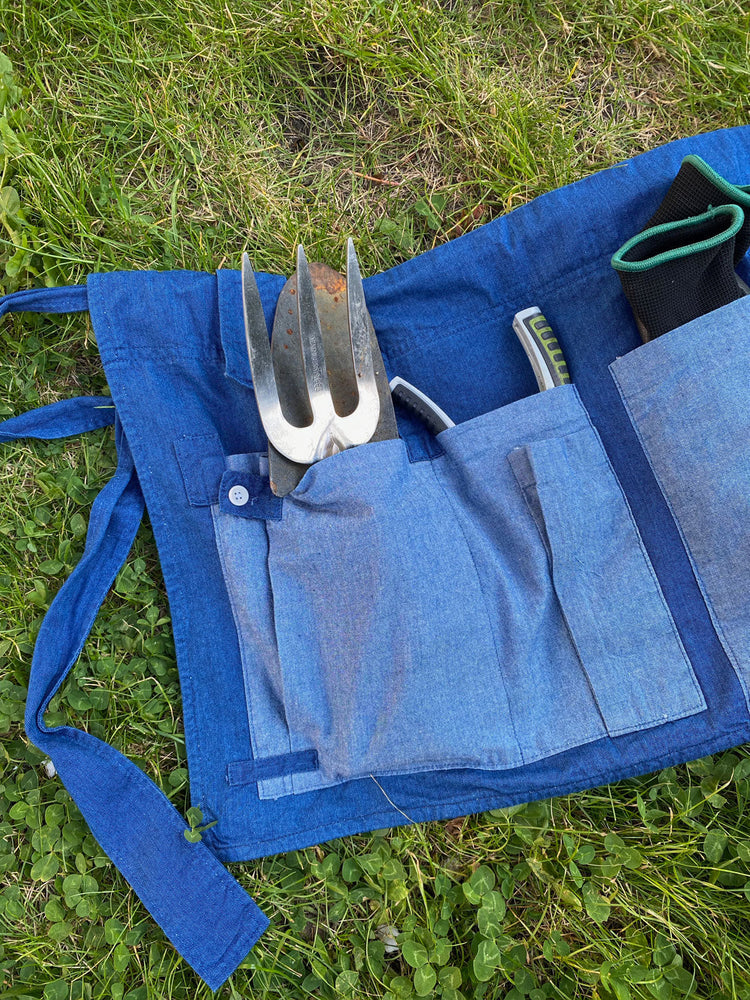 Sustainable Garden Tool Belt made from natural indigo dyed Japanese denim, by Saywood. Holds a garden trowel and fork, secateurs and gardening gloves, lying on the grass