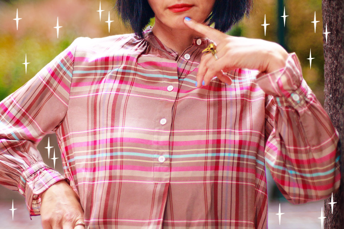 Close up of women wearing a pink tartan check blouse with gathered cuffs. She has her finger to her chin and is wearing gold rings. Stars surround her.