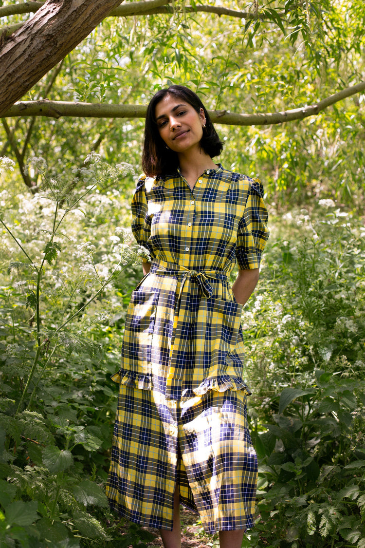 Model stands in a leafy green forest area, with her hands clasped behind her back and smiling. She wears Saywood's Rosa Puff Sleeve Dress in Yellow Check with the belt tied softly round her waist.