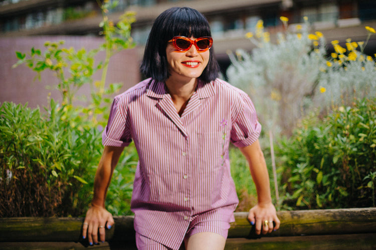 Women leans against a green rusty shipping container, with her arms folded. She wears Saywood's Zadie Boyfriends Shirt in Japanese Denim, with the waist belt tied.