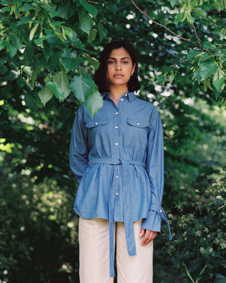 A woman stands amongst greenery and trees. She looks ahead, wearing Saywood's Japanese Denim Zadie Boyfriend Shirt with the belt tied round her waist, paired with beige trousers.