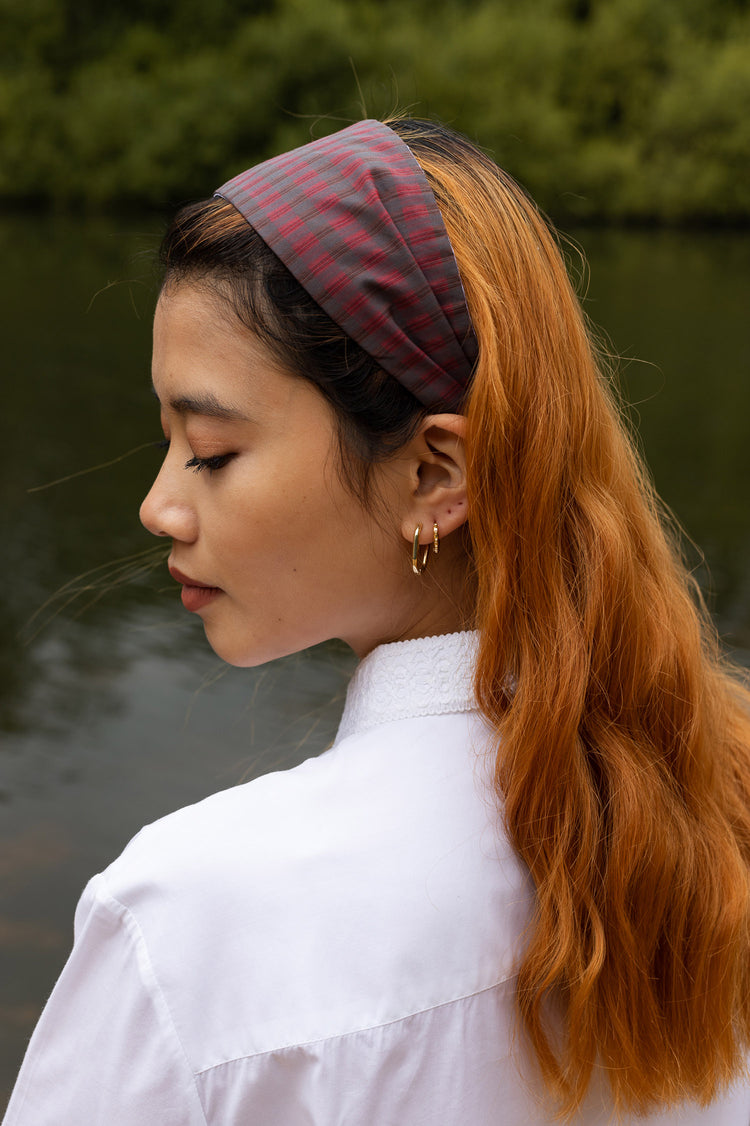 Model stands by a lake, wearing Saywood's sustainable Heidi Headband made from left over production cloth in red check deadstock cotton. Worn with the Edi Volume Sleeve White Shirt and gold earrings.