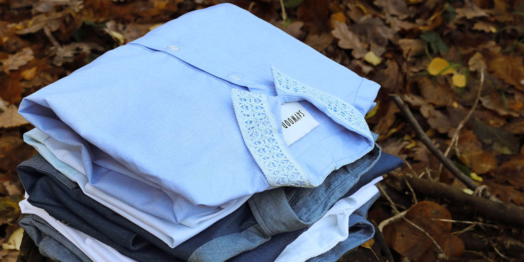 A pile of folded shirts, on a tree stump surrounded by fallen Autumnal leaves. The pale blue recycled cotton Edi Shirt with lace trim collar sits on the top of the pile.