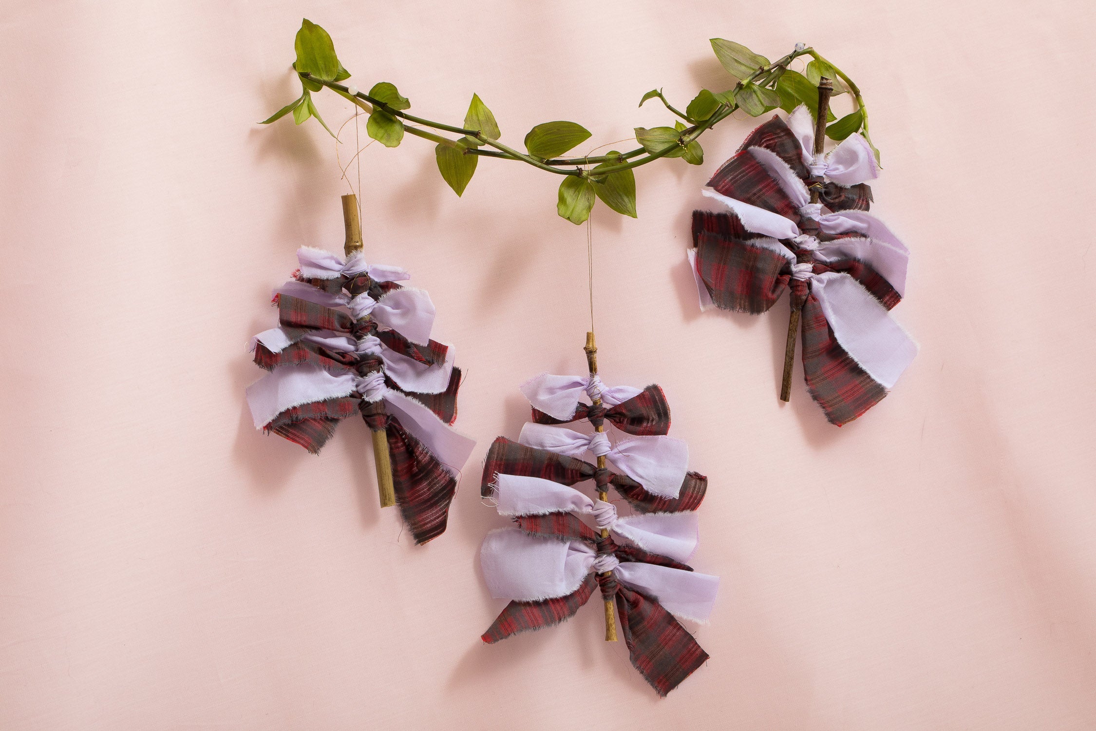 Three Christmas Tree Decorations in lilac and red check, sustainably made from natural materials, cotton surplus fabric and bamboo. Hanging on a piece of small green ivory against a pink packdrop.