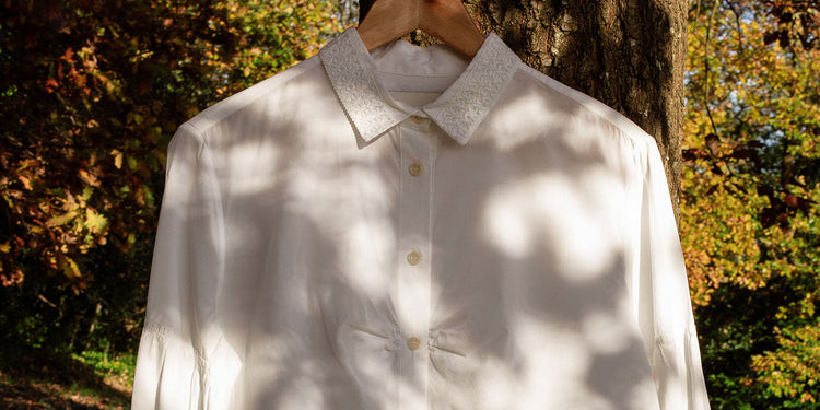 Saywood's white Edi Volume Sleeve Shirt with lace collar detail hangs on a wooden hanger in front of a tree, with leaves seen in the background. Light and shadow play on the front of the shirt, with shadows of the leaves visible.