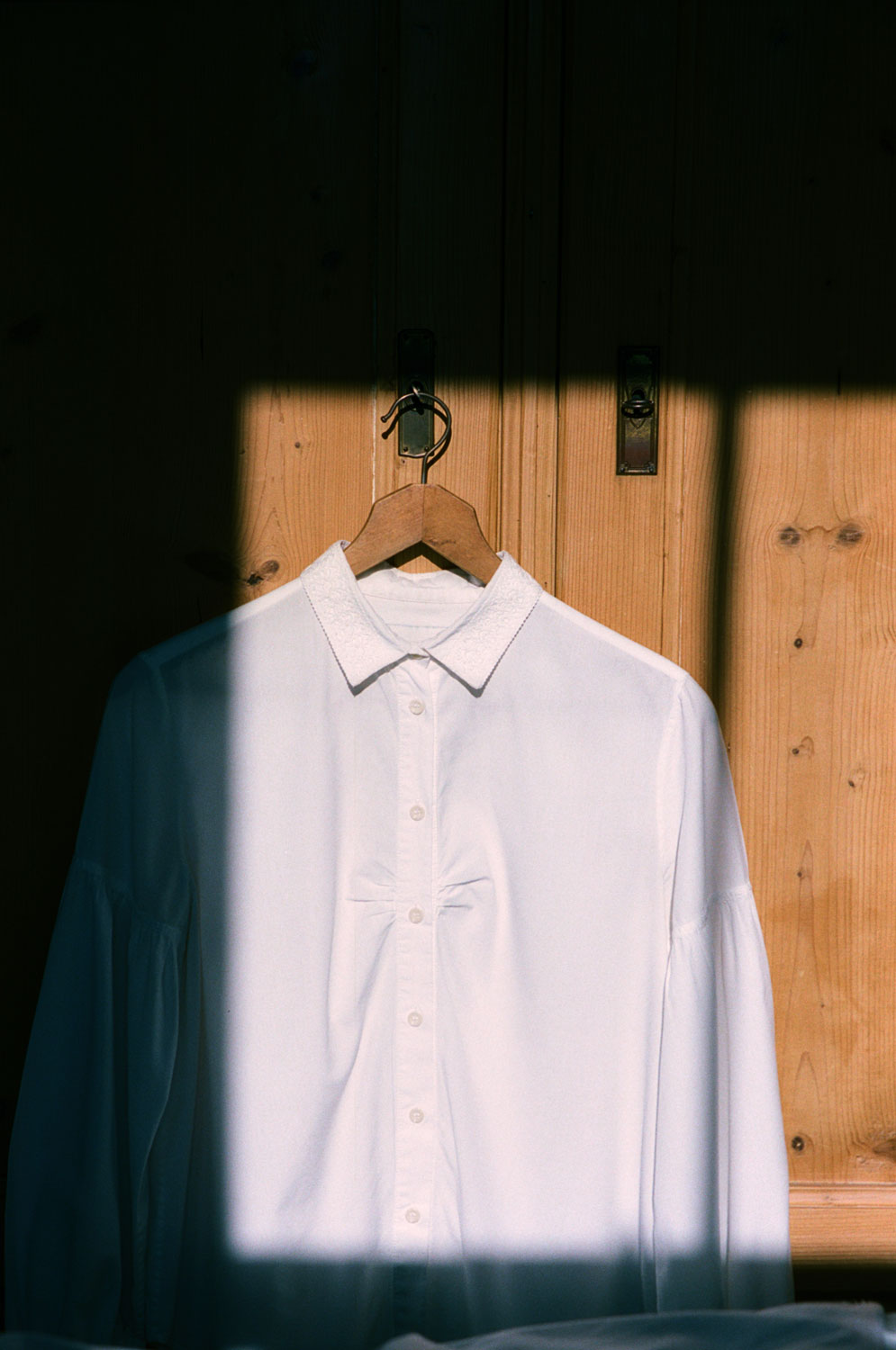 Saywood's Edi Volume Sleeve shirt with lace collar hangs on a wooden hanger on an old French pine wardrobe. The wardrobe is in shadow, with a square of sunlight directly falling on the shirt.