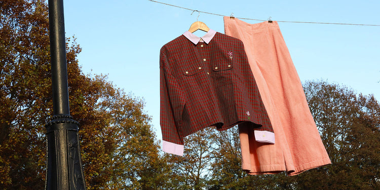 A shirt and trousers hang on a line from a black Victorian lamp post against a back drop of blue sky and trees. Saywood's Jules Utility Shirt in red mini check with contrast lilac collar and cuffs, with the pink corduroy Amelia Wide Leg Trousers