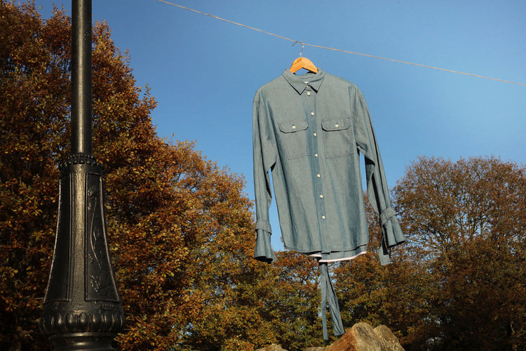 Saywood's womens Japanese Denim Zadie Boyfriend Shirt with safari styling details hangs from a line, from an old Victorian black lamp post. Blue sky and trees are seen in the background. 