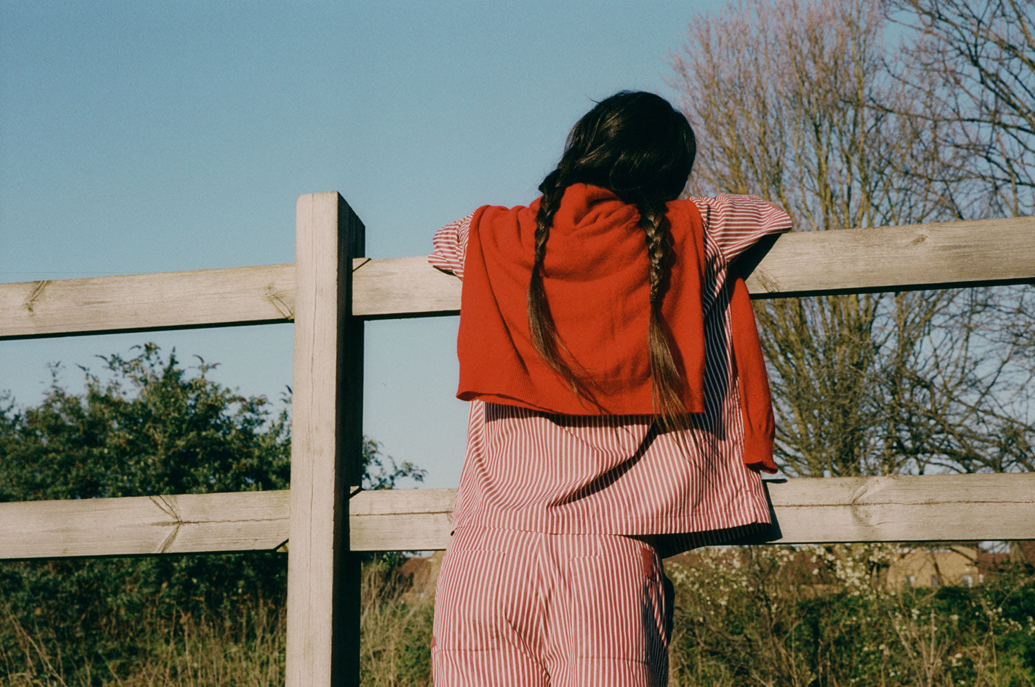 Woman with plaited pigtails leans on a wooden fence, overlooking a field. Her back is to the camera. She wears Saywood's Frances Unisex Cuban Shirt, Bessie A-line Shorts in berry red stripe as a matching set. A red cardigan is round her shoulders.