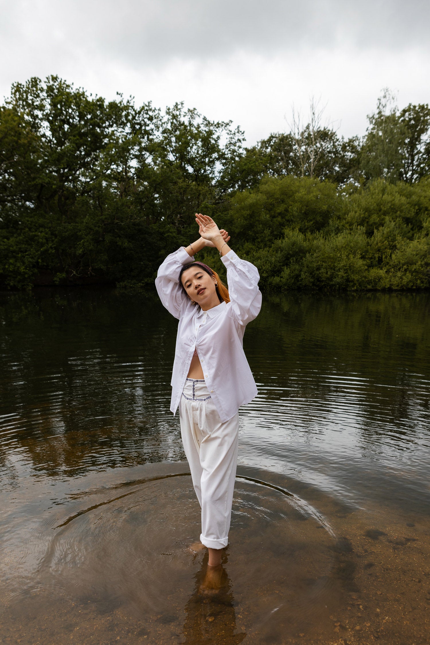 Model stands in a lake, surrounded by trees, with her hands above her head. She wears Saywood's Edi Volume Sleeve Shirt in white cotton bamboo, with the red check Heidi Headband in her hair. Her eyes are closed and she smiles softly.