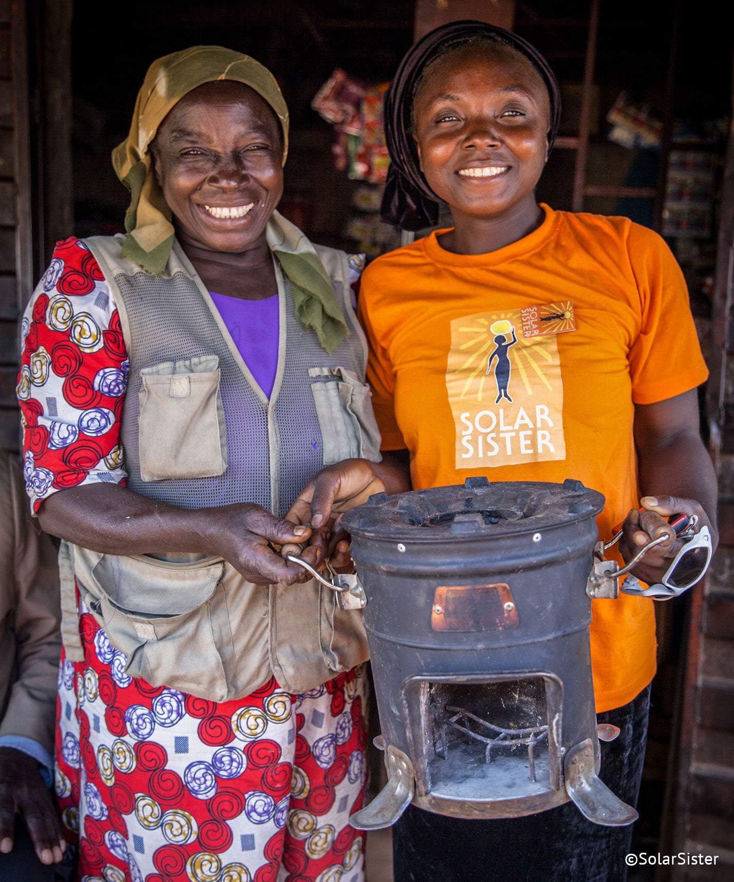 Two women stand together, smiling, holding and old coal stove. One woman wears an orange 'Solar Sister' tshirt