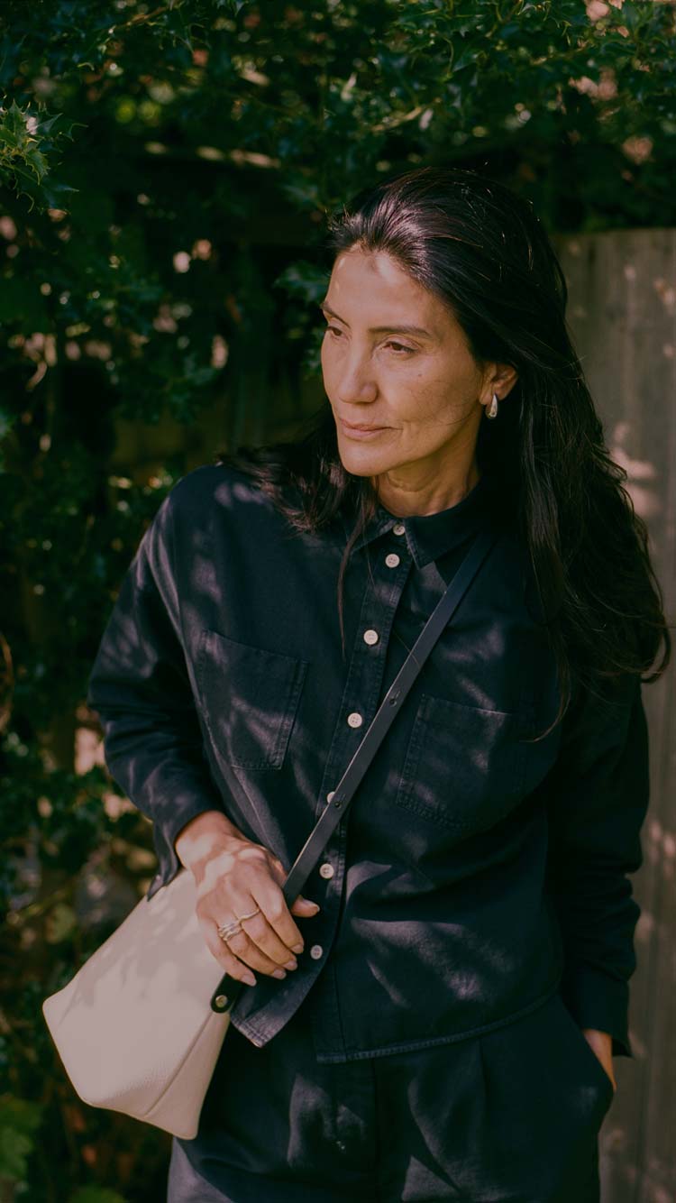 Lela Boxy Shirt in black Japanese denim with pale buttons, worn by a model carrying a beige crossbody bag in a leafy garden.