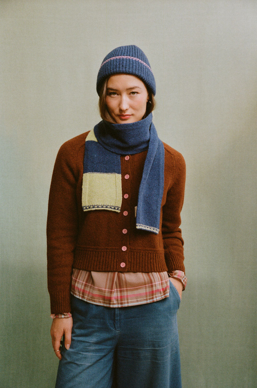 Front view of a model wearing an Atlantis blue beanie with pink trim, a rust cardigan, blue scarf and denim Amelia Trousers.
