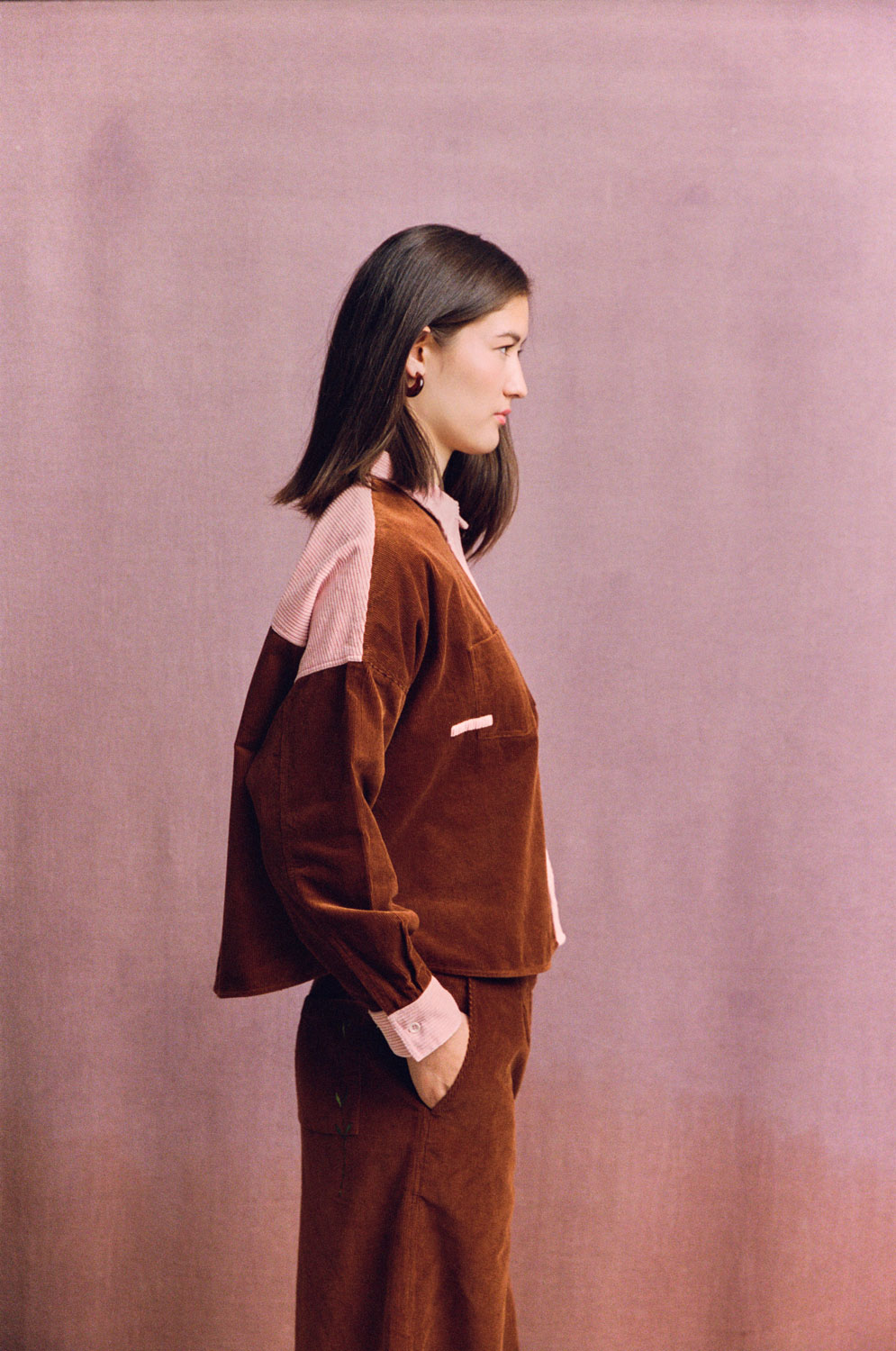 Lela caramel corduroy shirt shown from the side on a model, with pink contrast collar and cuffs against a muted pink backdrop.