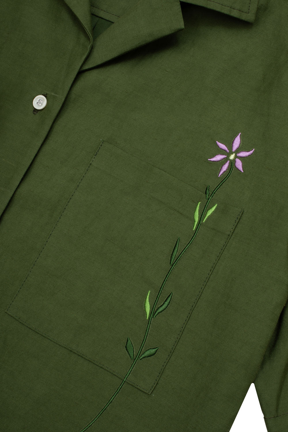 Close up of the Frances Unisex Cuban Shirt in olive green viscose cotton linen cloth. The embroidered flower signature to Saywood can be seen across the patch pocket on the chest.