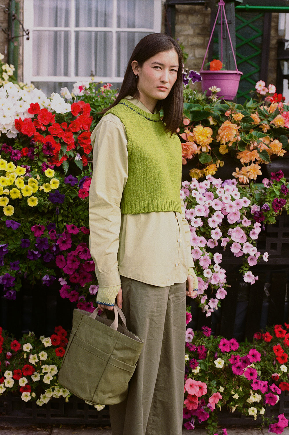 Ada green knitted vest layered over a cream shirt, styled with olive trousers on a model standing beside a flower display.