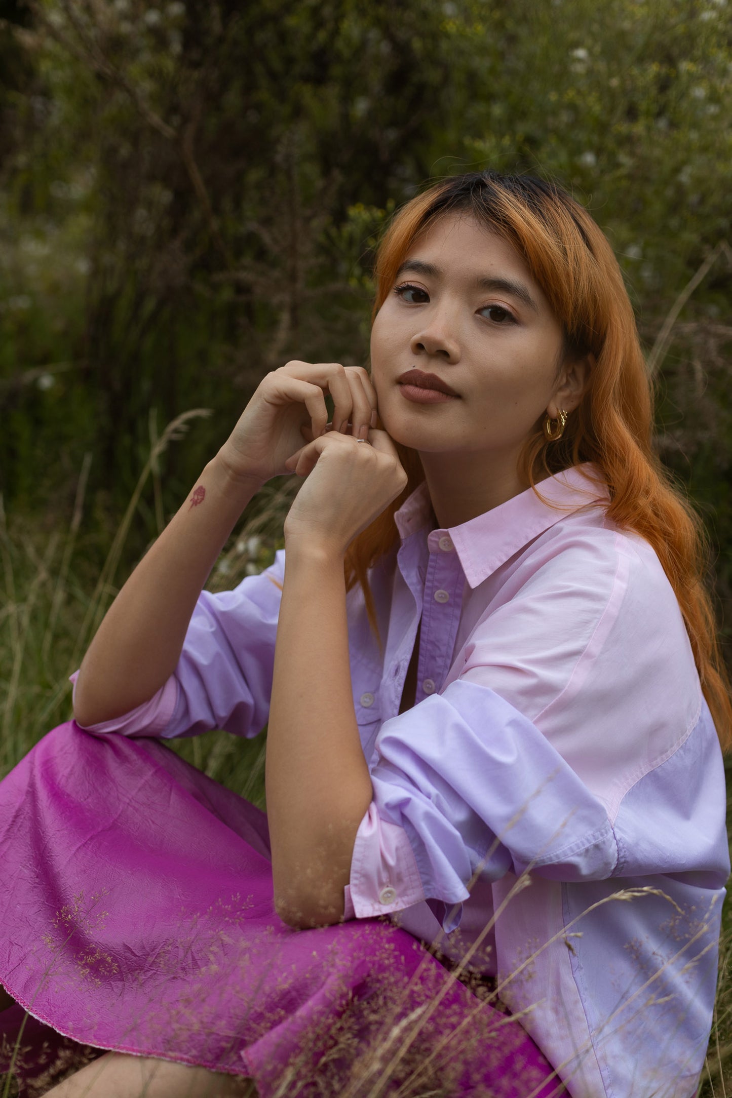 Model sits the grass surrounded by nature, looking at the camera. She wears Saywood's Jules Utility Shirt in lilac and pink colourblock cotton.