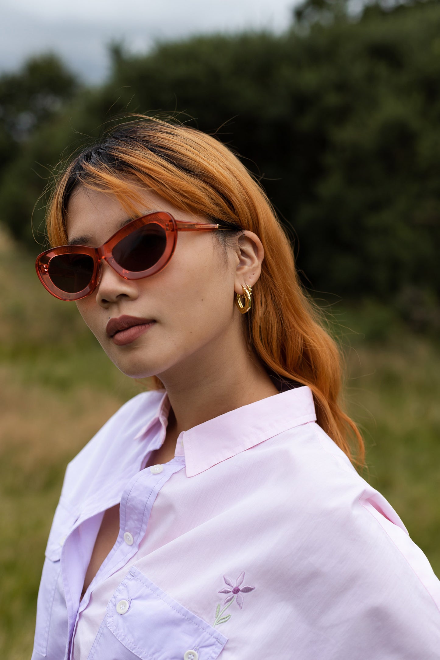 Close up of model standing in a field amongst nature, looking at the camera. She wears Saywood's Jules Utility Shirt in lilac and pink colourblock cotton, with the Saywood signature embroidered flower visible just above the patch utility pocket. Worn with red pink sunglasses.