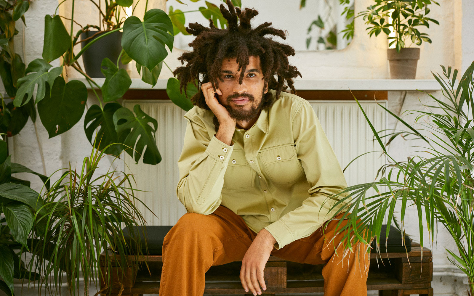 Model sits with his head resting on his hand, surrounded by house plants. He wears Saywood's Eddy Mens Utility Shirt in Olive with caramel trousers