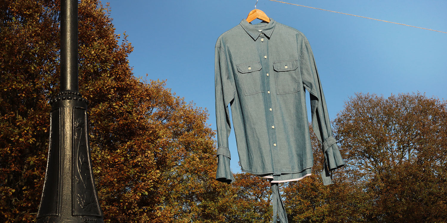 Women's Boyfriend Shirt in Japanese Denim - Saywood's Zadie Boyfriend Shirt in lightwash Japanese denim hangs from a line out in nature. The safari details and contrast internal hem bind details can be seen. Blue sky and trees are in the background, with an old Victorian lamp post on the left side of the image.